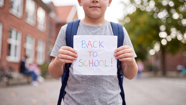 School boy holding back to school sign outside a school building with backpack for education season enrollment and classroom return promotion