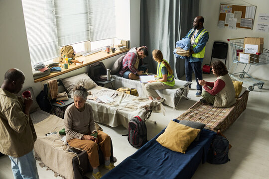 Wide angle view of diverse adults receiving shelter support, resting on cots, consulting volunteer staff in temporary housing center. Ideal for homelessness aid, crisis response campaigns