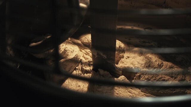 Close up fresh dough mixing process inside large spiral industrial mixer machine. View through protective grate. Preparing ingredients for baking bread. Artisan private bakery operations