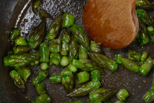 Chopped fresh green asparagus pieces are sauteed with oil in a dark frying pan with a wooden spoon stirrer