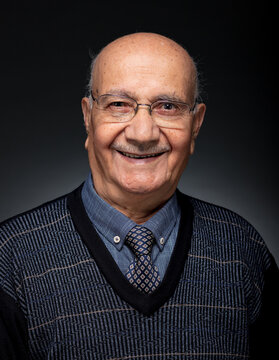 Portrait of a smiling senior man wearing glasses and a patterned sweater over a shirt and tie on a dark background.