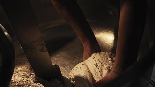Hands of female baker removing large piece of cooked dough from bowl of industrial mixer using metal scraper. Bakery kitchen preparation area. Process of removing dough for next stage of bread making