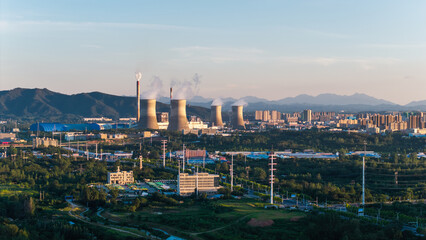 Fototapeta premium Thermal power plant with cooling towers emitting steam against urban skyline and distant mountains at dusk