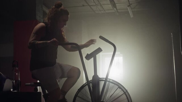 Young Caucasian woman mounting and riding air bike in dark gym. Footage ideal for fitness brands, gym advertisements, and healthy lifestyle promotional campaigns