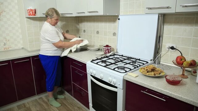 Older Caucasian woman washing dishes by hand in bright modern kitchen with gas stove, wiping plates with cloth near cabinet and countertop holding prepared food.