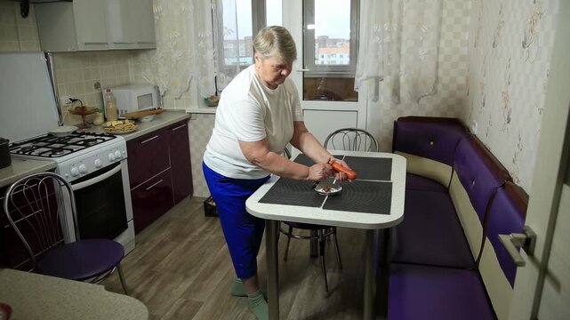 Older caucasian woman peeling carrot in small bowl on kitchen table, preparing food for cooking, wearing white t-shirt and blue pants, domestic scene of meal preparation