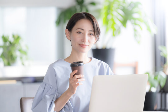 観葉植物のあるオシャレな明るいオフィスでノートパソコンで仕事をする日本人女性 オフィスワーク・ビジネスシーン　コーヒーを持つカメラ目線