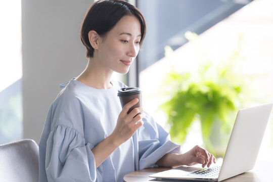 観葉植物のあるオシャレな明るいオフィスでノートパソコンで仕事をする日本人女性 オフィスワーク・ビジネスシーン　コーヒーを持つ横顔