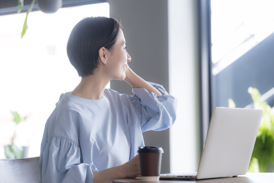 ノートパソコンとコーヒーを前に窓の外を眺める日本人女性 オフィスで休憩する働く女性のイメージの横顔