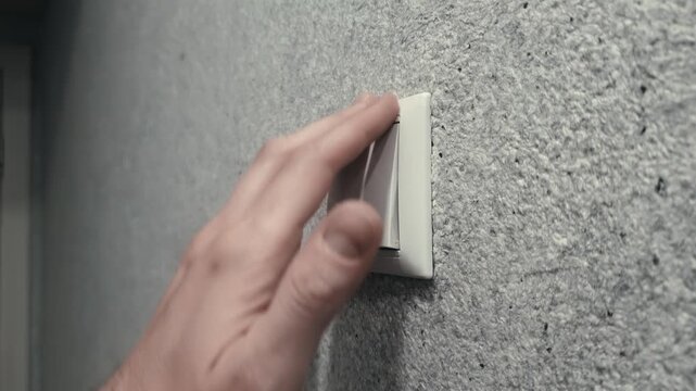 Male hand turns lights on and off. Activation of illumination in hallway of residence. Modern white light switch detail on textured grey wall. Close up view of electrical control panel