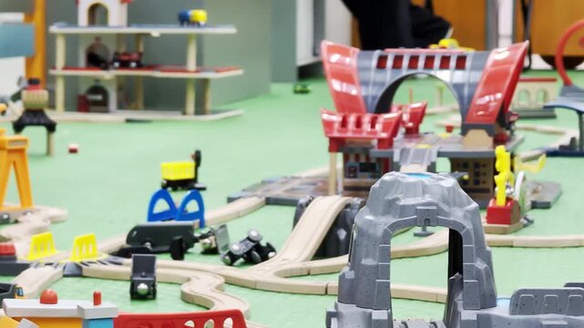 Extensive wooden toy train set layout on green mat. Network of curved and straight tracks, multiple bridges, tunnels, and stations. Colorful toy vehicles and accessories populate children play area.