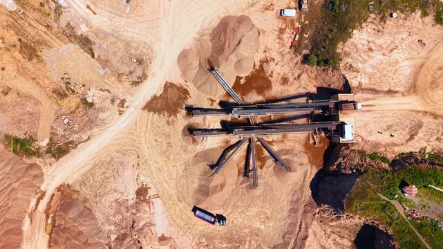 Sand quarry conveyor machinery operating with aggregate stockpiles. Aerial perspective showing industrial equipment, earth heaps, trucks on dry ground in daylight.