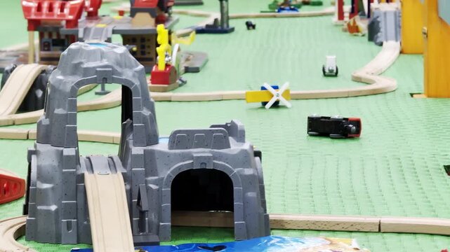 Colorful collection of children toy trains, cars, and buildings creating an elaborate play scene on a soft green mat in a bright indoor playroom.