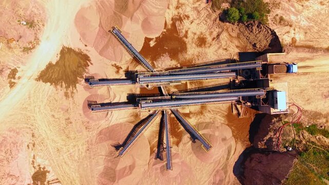 Aerial view of conveyor belts processing gravel in a quarry. Overhead perspective of industrial equipment, sandy terrain, brown soil and stone stockpiles in bright sunlight.