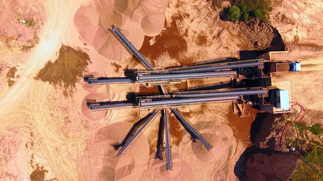 Conveyor belt system sorting aggregate materials in a quarry. Vertical top view of industrial transport machinery, gravel piles, sandy dirt, trucks and bright lighting.