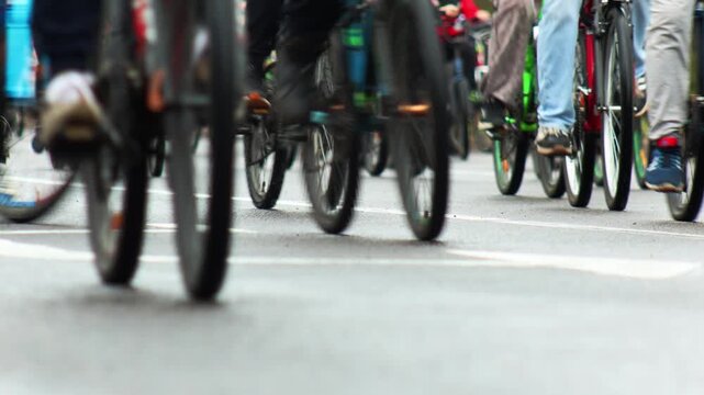 Group of cyclists participate in a bike ride on asphalt road during daytime, showcasing motion blur of numerous bicycle wheels and legs of people, creating a dynamic sense of movement.