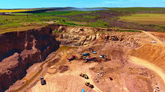 Wide aerial panorama of open pit quarry and solar farm. High perspective showing excavated terrain, machinery, green hills, photovoltaic modules and blue sky daylight.
