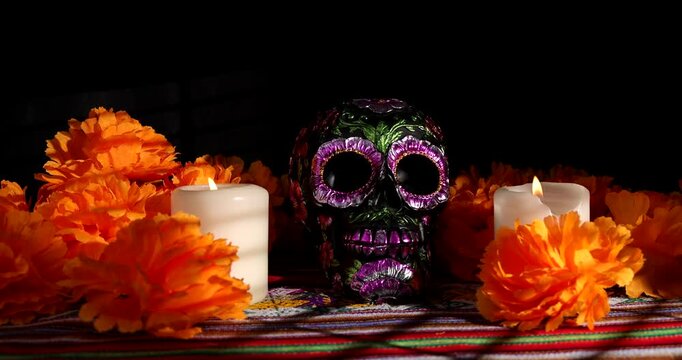 A white candle is placed carefully on a traditional Mexican altar beside a black metallic painted skull and red flowers against dark background.