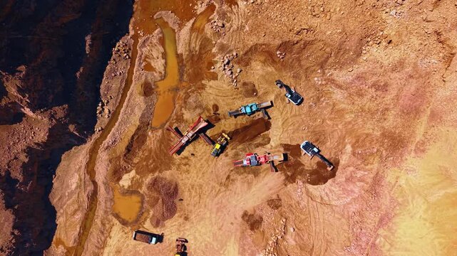 Heavy excavators operating in open pit stone quarry. Top down aerial photo capturing crushing machinery, dump trucks, muddy puddles on reddish soil.