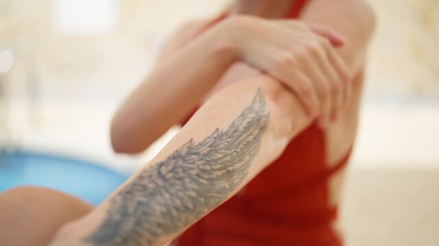 Young woman in a red swimsuit carefully applying moisturizing sunscreen on her arm with a delicate wing tattoo, preparing for a relaxing day of sunbathing by the swimming pool