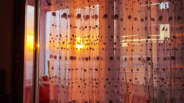 Golden hour sunset light streams through translucent decorative beaded curtain hanging in front of balcony window, casting warm atmospheric glow across interior space, revealing residential buildings
