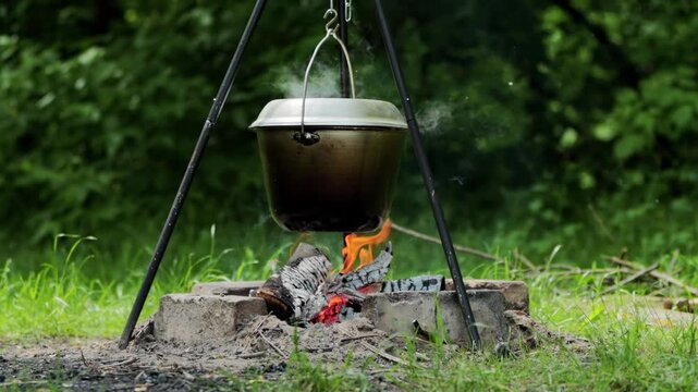 Metal cooking pot with lid hanging from metal tripod stand boiling over open campfire with vibrant flames and rising smoke for outdoor meal preparation slow motion