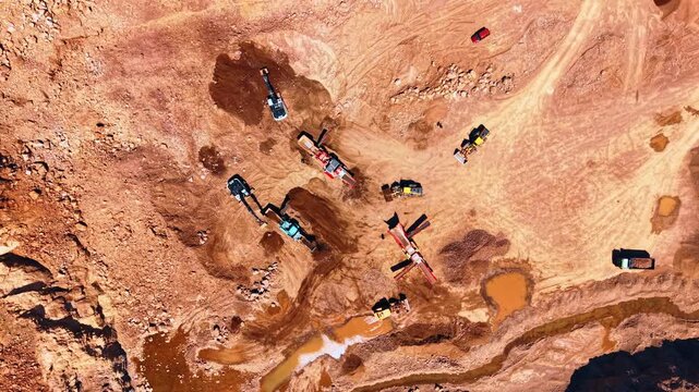 Top view of excavators and screening machines in open gravel pit. Heavy machinery operates on reddish brown soil near mud puddles and dirt paths in an active industrial site.