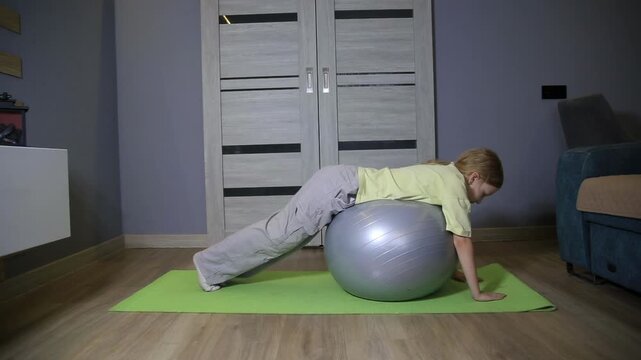Young caucasian girl performing stability ball exercises at home living room, fitness workout for core strength and balance development, active child exercising indoors on yoga mat