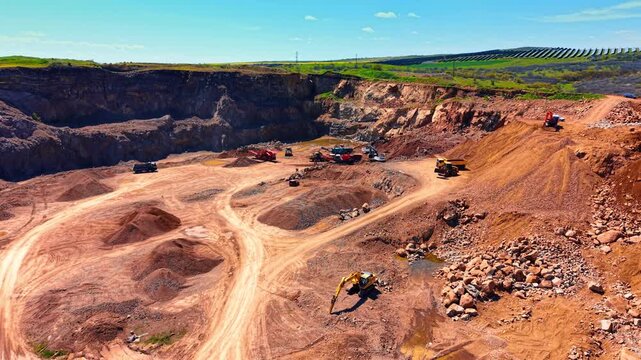 Open pit quarry site with heavy machinery and stone piles. Wide aerial panorama of industrial mining landscape with trucks and red earth under blue sky.