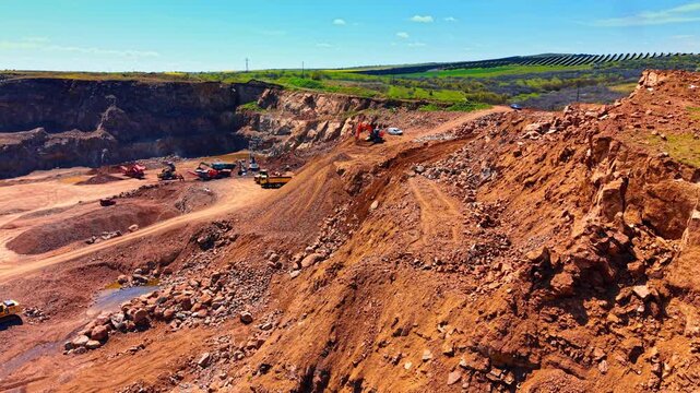 Open stone quarry with terraced rocky slopes and solar farm horizon. High angle perspective shows red earth slopes and machinery with green fields and solar panels under blue sky.