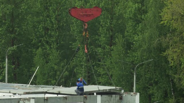 Construction workers guiding massive concrete slab placement on unfinished building structure with powerful red crane hook against background of green trees daytime