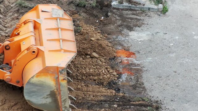 Heavy equipment excavator bucket dumping pile of earth and dirt from construction site next to asphalt road, revealing wet ground and orange liquid, slow motion