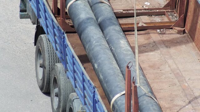 Truck with blue side rails transporting two large dark pipes securely fastened with straps on a rusty flatbed, viewed from above, moving on asphalt road, slow mo