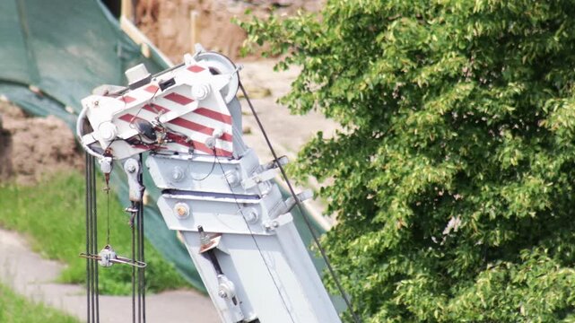 Close up view of top part of construction crane boom with pulley system and steel cables, moving slightly against green construction netting and lush green tree foliage, daylight scene, slow mo