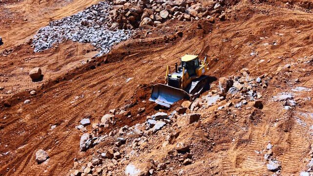 Yellow bulldozer moving rocks on dirt slope at quarry site. Heavy crawler dozer clears red rocky terrain in daylight captured from high angle.