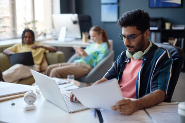 Fototapeta premium Young man working on laptop and reviewing documents in shared office while colleagues using devices in background, representing teamwork and productivity. For startup marketing or workplace content