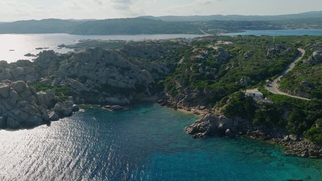 Drone view of a secluded cove at Capo Testa near Santa Teresa Gallura, featuring granite cliffs, turquoise Mediterranean water, green vegetation and a winding coastal road.