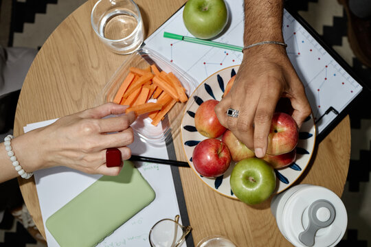 Overhead shot of adult coworkers sharing apples and carrot sticks over work documents, promoting healthy office snacking and wellness. Ideal for workplace nutrition or wellbeing campaigns