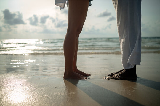 Interracial couple standing close on a sandy beach, focusing on bare feet and a moment of intimacy