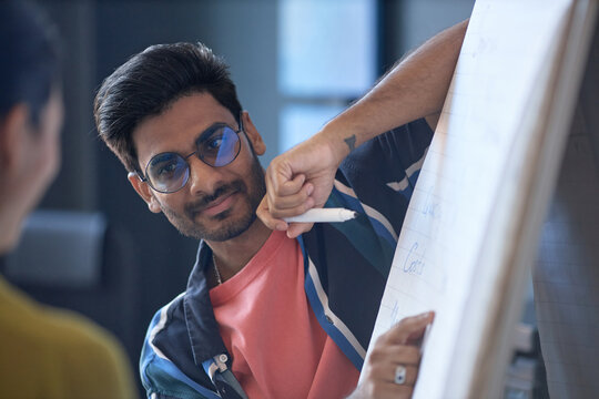 Young adult Indian man presenting ideas on flip chart during meeting, explaining strategy to colleague. Useful for business training, teamwork, coaching, workplace communication