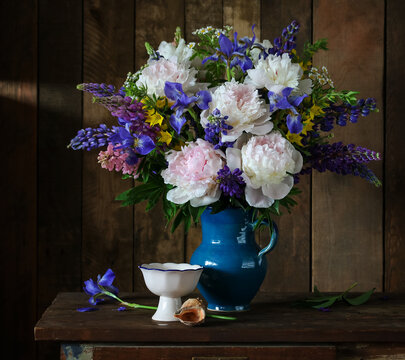 Lush bouquet of pink peonies, blue irises, and lupines in a ceramic pitcher on a wooden table
