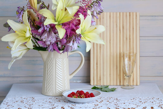 Floral bouquet with lilies in pitcher sits beside fresh raspberries on a decorated table. Summer still life, home decoration, and country living concept.