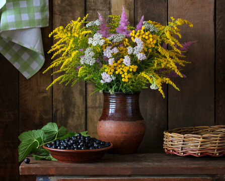 Wildflower bouquet in a ceramic jug stands on a wooden table beside a bowl of fresh black currant. Rustic still life and summer countryside concept.
