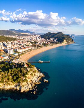 Aerial view of a coastal city, showcasing a long sandy beach, turquoise water, and a scenic mountainous background