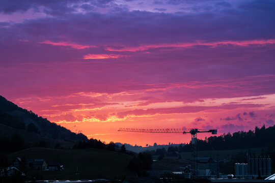 Tower crane silhouetted against a spectacular purple and orange sunset over a peaceful rural landscape, with dramatic clouds creating a vibrant twilight sky and highlighting the harmony between modern