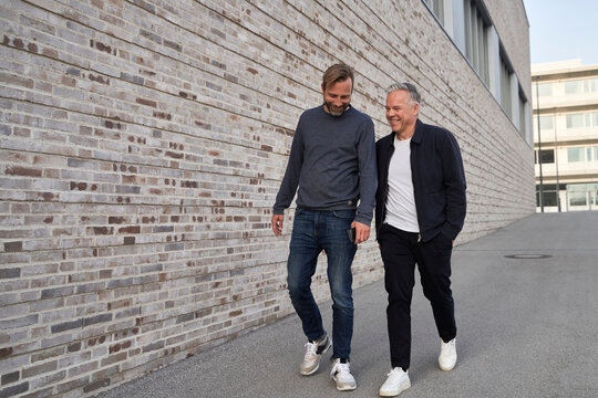 Smiling male professionals walking on footpath by brick wall