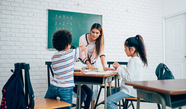 Happy school teacher and students having interesting engaging activities in class. Little children sitting around classroom table, talking and playing fun games together