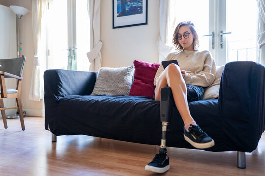 Young woman with leg prosthesis sitting on couch at home reading e-book