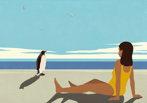 Girl in bathing suit relaxing on sunny summer beach, watching penguin standing nearby
