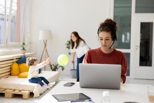 Young woman working from home while nanny is playing with daughter